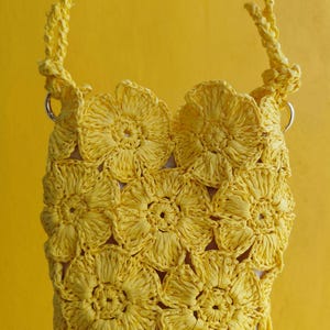 May include: A bright yellow crochet bag constructed from multiple joined floral motifs. The bag features a textured, open-work design with a matching yellow braided strap attached to silver-toned metal rings on each side of the top opening.