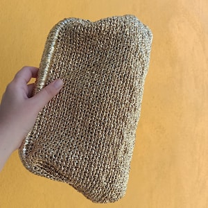Op de afbeelding: Een goudkleurige clutch met een geweven textuur. De rechthoekige tas wordt door een hand tegen een gele achtergrond gehouden. De clutch heeft een metallic glans en een eenvoudig, elegant ontwerp.