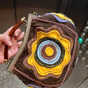 Puede incluir: Un bolso de ganchillo con un patrón circular de estrella multicolor en tonos marrón, amarillo y azul claro. El bolso tiene un marco metálico con una textura grabada ornamentada y un asa curva con aspecto de carey marrón oscuro.