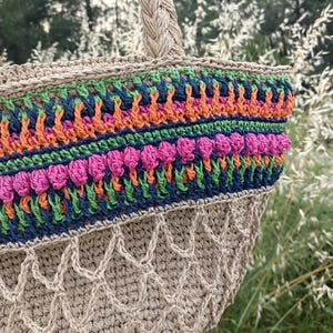 Peut inclure: Un sac cabas en crochet beige doté d'une anse tressée et d'une bande décorative supérieure avec des rayures horizontales bleu marine, vert vif, orange et rose fuchsia. La partie inférieure du sac présente un motif en filet ajouré.