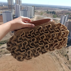 May include: A tan crocheted clutch with a brown leather handle. The clutch has a ruffled, textured design and a rectangular shape. The background shows a cityscape with tall buildings under a clear, sunny sky.