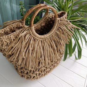 May include: A natural-coloured woven straw tote bag with bamboo handles. The bag has a fringed design around the top. The handles are made of curved bamboo.