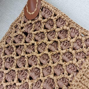 May include: Close-up of a woven straw tote bag with a brown leather handle. The bag features a geometric pattern with alternating light and dark brown woven sections. The handle is attached with a decorative leather piece.