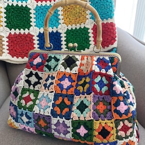 Peut inclure: Un sac à main crocheté multicolore avec une poignée en bambou. Le sac est composé de différents carrés aux tons de rouge, bleu, vert, orange et violet. Un coussin crocheté assorti est en arrière-plan. Le sac a une fermeture à fermoir en métal.