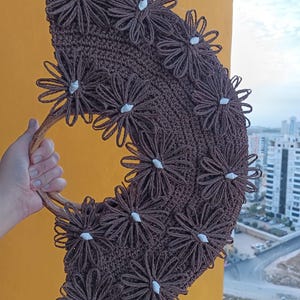 May include: A brown crocheted handbag with a wooden handle. The bag is decorated with numerous brown yarn flowers, each with a small white center. The bag is held up against a yellow background with a cityscape visible in the distance.