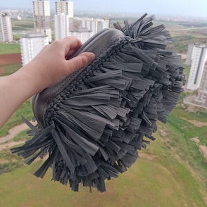 Könnte beinhalten: Eine graue Clutch mit Fransen-Design. Die Clutch wird vor einem Hintergrund aus Gebäuden und grünem Gras gehalten. Die Fransen bestehen aus langen, dünnen Materialstreifen, und die Clutch hat einen geflochtenen Rand.