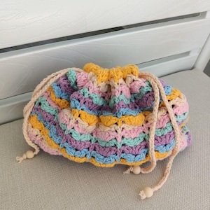 May include: A handmade crochet bag in a shell shape, featuring a drawstring closure. The bag showcases a colorful striped pattern with shades of yellow, pink, blue, and purple. The drawstring is a light beige color with wooden beads at the ends.