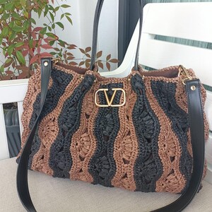May include: A large, woven tote bag with alternating vertical stripes of black and brown. The bag features black leather handles and a detachable shoulder strap. A gold-colored "V" logo is centered on the front.