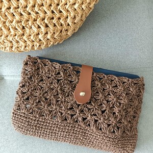 May include: A brown crochet clutch with a floral pattern and a leather strap closure. A woven straw tote bag is partially visible in the background. The clutch is a stylish accessory.