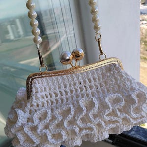 Könnte beinhalten: Eine weiße gehäkelte Handtasche mit gerüschter Textur und einem goldfarbenen Metallrahmen mit einem Verschluss aus zwei Perlen. Die Tasche hat einen abnehmbaren Henkel aus einer Reihe weißer Kunstperlen, befestigt mit goldfarbenen Metallclips.