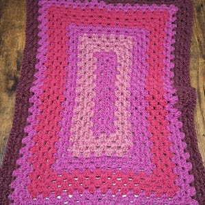 May include: A rectangular crochet blanket featuring a concentric granny square pattern. The design consists of alternating bands of yarn in shades of dark maroon, vibrant magenta, and soft dusty rose, creating a layered, multi-colored effect.