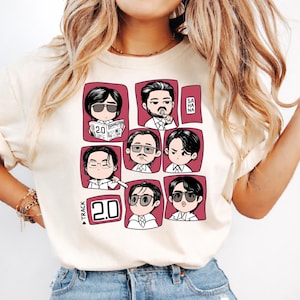 以下が含まれることがあります： サングラスをかけた漫画の肖像画のグリッドと「2.0」と「TRACK」の文字が特徴のクリーム色のTシャツ。肖像画はマルーンの背景に配置されています。シャツは柔らかい素材で作られています。