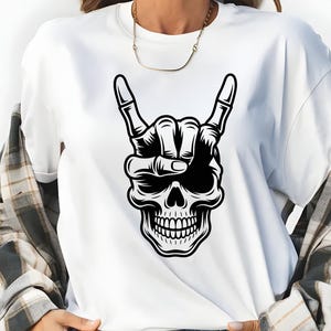 May include: A white short-sleeved t-shirt featuring a black graphic print of a human skull with a skeletal hand making a rock and roll hand gesture above its head. The shirt is worn with a grey and white plaid button-down shirt and blue jeans.