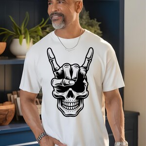 May include: A white short-sleeved t-shirt featuring a large black and white graphic print of a human skull making a rock and roll hand gesture. The skull is positioned centrally on the chest area of the shirt, displaying detailed bone structure.