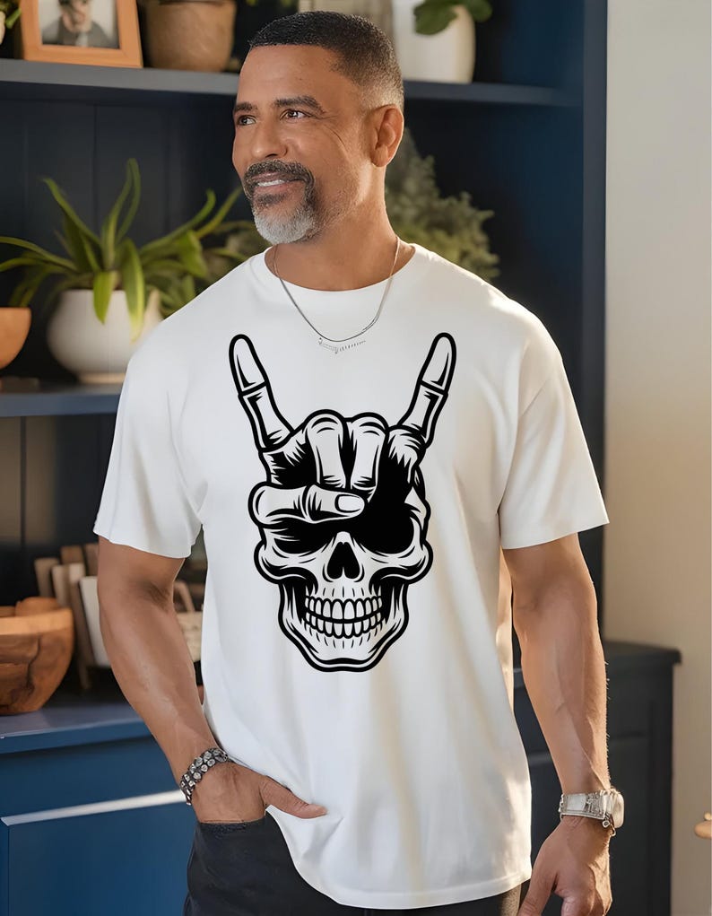 May include: A white short-sleeved t-shirt featuring a large black and white graphic print of a human skull making a rock and roll hand gesture. The skull is positioned centrally on the chest area of the shirt, displaying detailed bone structure.