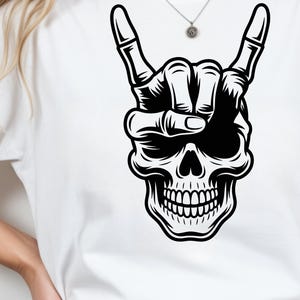 May include: A white t-shirt featuring a bold black graphic print of a human skull. The skull is depicted with a hand positioned above it, forming a rock and roll hand gesture with the index and pinky fingers extended upward.