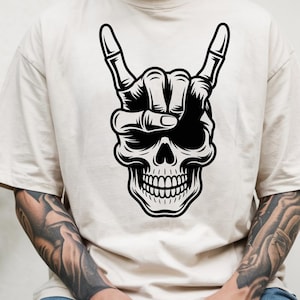 May include: A beige short-sleeved t-shirt featuring a large black and white graphic print on the chest. The design depicts a human skull with a skeletal hand positioned over the forehead, displaying the rock and roll hand gesture.
