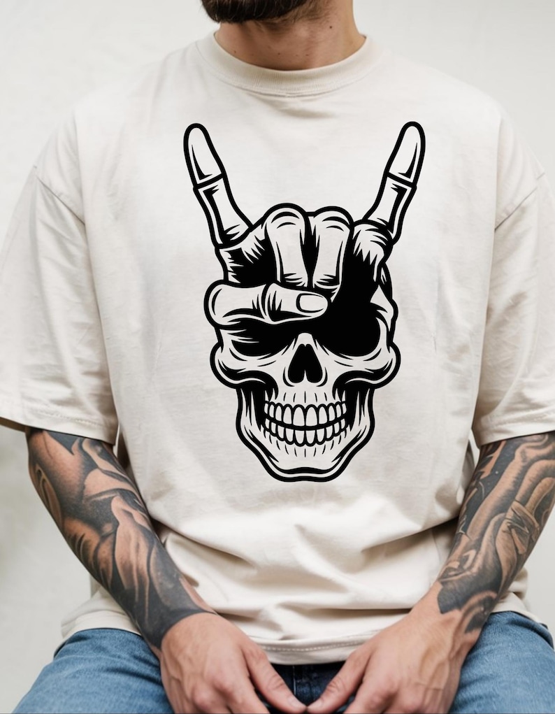 May include: A beige short-sleeved t-shirt featuring a large black and white graphic print on the chest. The design depicts a human skull with a skeletal hand positioned over the forehead, displaying the rock and roll hand gesture.