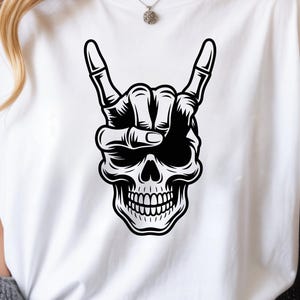 May include: A white t-shirt featuring a black graphic print of a human skull with a skeletal hand positioned above it, making a rock and roll hand gesture. The design is centered on the chest, showcasing detailed line work and shading.