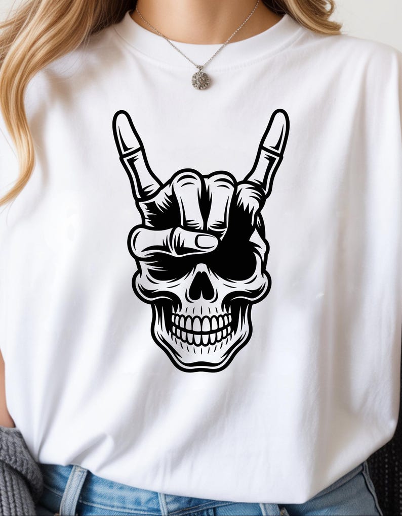 May include: A white t-shirt featuring a black graphic print of a human skull with a skeletal hand positioned above it, making a rock and roll hand gesture. The design is centered on the chest, showcasing detailed line work and shading.
