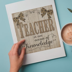 May include: A white framed print with the text "TEACHER is an ocean of Knowledge" in a rustic font, adorned with leopard print bows. A cup of coffee and a sprig of leaves sit on a light blue surface.