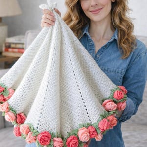 Pode incluir: Um cobertor de bebê de crochê creme com uma borda floral. O cobertor é levantado, mostrando sua forma triangular e o trabalho de crochê detalhado. A borda apresenta rosas rosa e vermelhas com folhas verdes.