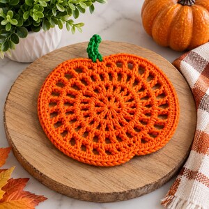 May include: A round, orange crochet pumpkin coaster featuring an openwork, radial stitch pattern and a small, solid green stem at the top. The piece is crafted from yarn and displays a textured, circular design that mimics the shape of a pumpkin.