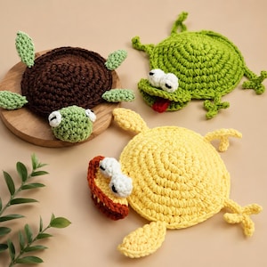 Puede incluir: Tres posavasos de ganchillo con forma de tortuga marrón, rana verde con lengua roja y pato amarillo con pico naranja. Cada posavasos presenta ojos redondos blancos y una construcción de hilo texturizado con formas animales.