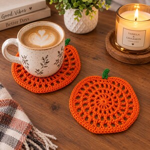 May include: Two round orange crochet coasters shaped like pumpkins with small green stems. One coaster sits under a speckled ceramic mug filled with latte art, while the other lies flat. The design features an openwork pattern of radiating stitches.