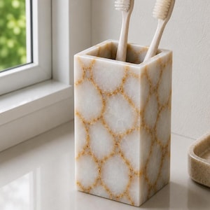 Puede incluir: Un portacepillos rectangular de piedra blanca con un patrón dorado en forma de telaraña. Dos cepillos de dientes beige están dentro del soporte. El soporte está sobre una superficie blanca cerca de una ventana.