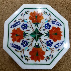 May include: An octagonal white marble box with a floral inlay design. The design features orange flowers, blue accents, and green leaves. The box is edged with a green border.