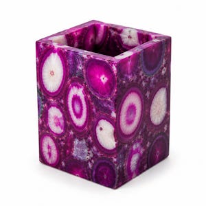 May include: A square, decorative container made of polished, purple agate. The container features a pattern of concentric circles in shades of white, pink, and purple. The interior is hollow, suggesting it could be used as a pen holder or small vase.