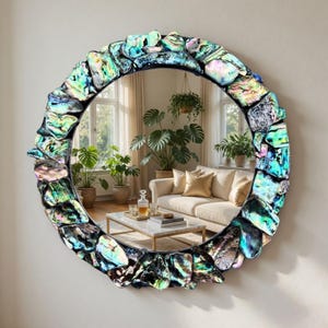 May include: Circular wall mirror featuring a frame adorned with iridescent, multi-coloured stones. The mirror has a black frame and reflects a room with plants and a sofa. The stones display hues of blue, green, and purple.