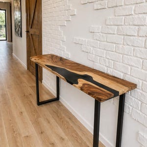 Puede incluir: Una consola de madera con un diseño de río de resina epoxi negra. La mesa tiene una tapa rectangular con vetas de madera natural y un centro de resina epoxi negra. Se apoya en patas de metal negro. La mesa mide aproximadamente 152 cm de largo.