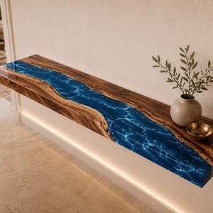 Op de afbeelding: Een zwevende wandplank van natuurlijk bruin hout met een centrale rivierachtige inleg van diepblauwe hars met witte wervelende patronen. Een kleine keramische vaas met groene bladeren en een kleine gouden kom staan op het houten oppervlak.