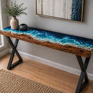 May include: A long, wooden console table with a unique ocean-themed epoxy resin top. The top features a realistic depiction of waves and pebbles. The table has black metal X-shaped legs. The table is approximately 60 inches long.