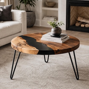 Peut inclure: Une table basse ronde présentant une surface en bois naturel avec une incrustation centrale en résine noire. La table est soutenue par trois pieds fins en épingle à cheveux en métal noir et supporte deux livres empilés et une petite plante.
