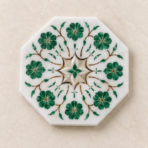 Op de afbeelding: Een achthoekige onderzetter van wit marmer met een ingewikkeld inlegontwerp van donkergroene bloemmotieven. Het patroon bestaat uit acht symmetrische bloemen verbonden door dunne bruine stengels en bladeren rond een witte ster.