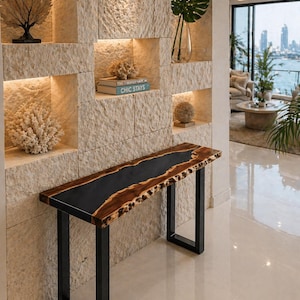 Pode incluir: Uma mesa de consola de madeira com um design de rio em resina epóxi preta e borda natural. A mesa tem pernas de metal preto. O fundo apresenta uma parede de pedra com prateleiras embutidas exibindo itens decorativos e o texto "CHIC STAYS".