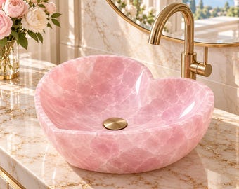 Puede incluir: Un lavabo de sobreponer en forma de corazón hecho de piedra rosa translúcida con acabado pulido. El cuenco presenta una tapa de desagüe circular dorada en el centro y se combina con un grifo curvo dorado a juego montado en la encimera.