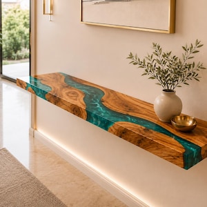 Op de afbeelding: Een zwevende wandplank gemaakt van natuurlijke houten platen met een centrale rivierachtige inleg van doorschijnende blauwgroene hars. De plank heeft een gladde, gepolijste afwerking die de houtnerf benadrukt en is horizontaal gemonteerd.