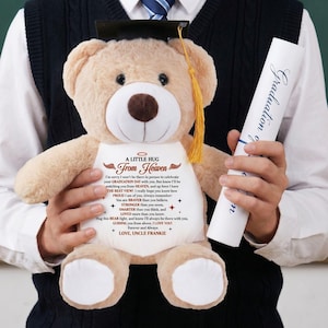 May include: A tan teddy bear wearing a graduation cap and holding a rolled-up diploma. The bear has a white shirt with the text "A LITTLE HUG From Heaven" and a message about graduation. A thoughtful gift for a graduate.