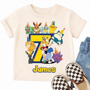 Puede incluir: Una camiseta de manga corta color crema con un número siete grande de color amarillo y borde azul. El diseño incluye varios personajes de dibujos animados con gorros de fiesta coloridos, un personaje humano y el nombre James en amarillo.