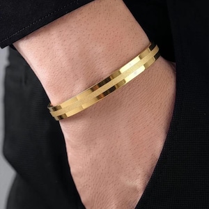 Può includere: Un bracciale rigido in metallo color oro caratterizzato da una finitura lucida sui bordi e una fascia centrale testurizzata con finitura opaca. Il bracciale presenta un design rigido e aperto che avvolge il polso, con un'estetica metallica.