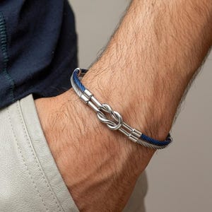 Op de afbeelding: Een armband met een ontwerp van twee strengen, bestaande uit een zilverkleurige metalen kabel en een donkerblauwe kabel. De twee strengen zijn in het midden verbonden door een gepolijst zilverkleurig metalen knoopelement met bijpassende eindkappen.