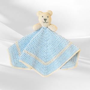 May include: A handmade crochet baby lovey featuring a light blue square blanket with a cream-colored border. A cream-colored teddy bear head is attached to the top of the blanket. The lovey is made of soft yarn.