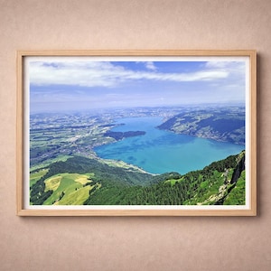 May include: A framed landscape print showcasing a vibrant aerial view of a lake surrounded by lush green mountains and fields. The water is a striking turquoise, contrasting with the blue sky and fluffy white clouds. The frame is made of light-colored wood.
