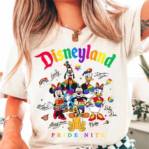Puede incluir: Camiseta blanca con la palabra "Disneyland" en colores arco&iacute;ris, junto con personajes de dibujos animados como Mickey y Minnie Mouse, Goofy, Donald y Daisy Duck y Pluto. El texto "Pride Nite" est&aacute; en la parte inferior.