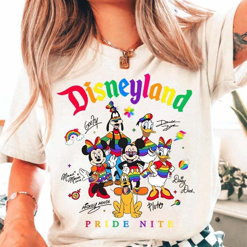 Puede incluir: Camiseta blanca con la palabra "Disneyland" en colores arco&iacute;ris, junto con personajes de dibujos animados como Mickey y Minnie Mouse, Goofy, Donald y Daisy Duck y Pluto. El texto "Pride Nite" est&aacute; en la parte inferior.