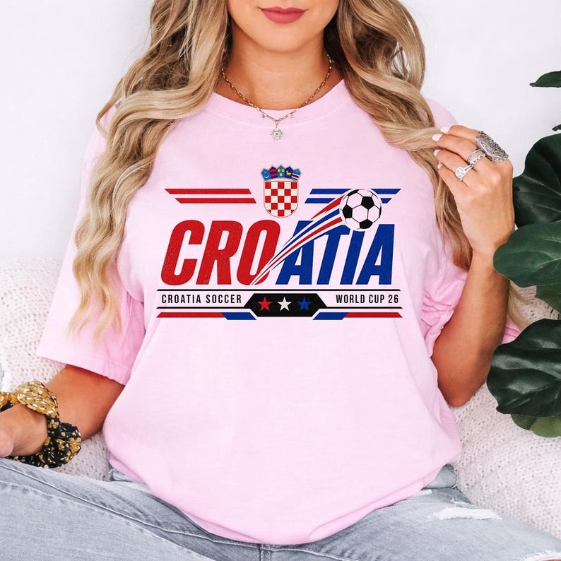 May include: A light pink short-sleeve t-shirt featuring a graphic design with the text CROATIA in red and blue letters, a soccer ball, the Croatian coat of arms, and the text CROATIA SOCCER WORLD CUP 26 with three stars in red, white, and blue.